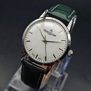Pu&ograve; includere: Un orologio Jaeger-LeCoultre Club argentato con quadrante bianco e cinturino in pelle nera. L'orologio ha un cinturino in pelle verde e le parole "SWISS MADE" sono visibili sul quadrante. L'orologio &egrave; esposto su un supporto trasparente.