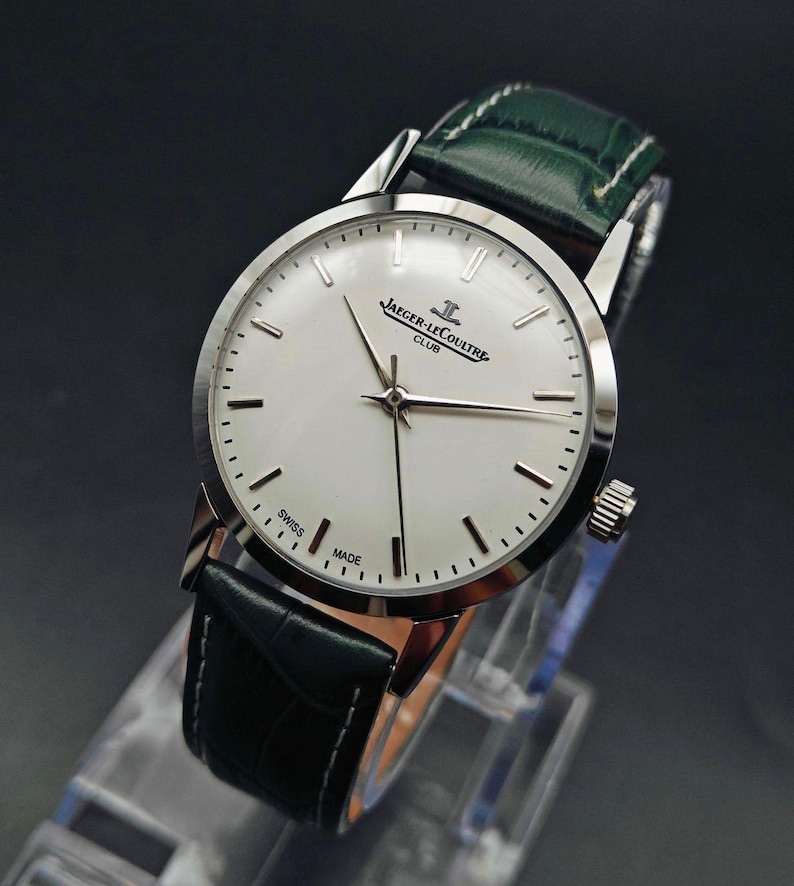Pu&ograve; includere: Un orologio Jaeger-LeCoultre Club argentato con quadrante bianco e cinturino in pelle nera. L'orologio ha un cinturino in pelle verde e le parole "SWISS MADE" sono visibili sul quadrante. L'orologio &egrave; esposto su un supporto trasparente.