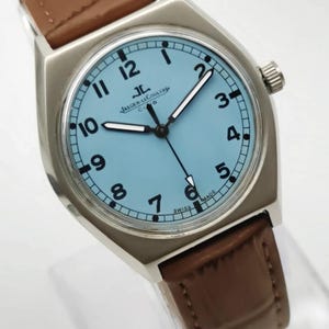K&ouml;nnte beinhalten: Eine silberfarbene Armbanduhr mit hellblauem Zifferblatt und schwarzen Ziffern. Die Uhr hat ein braunes Lederarmband und den Text "JAEGER-LECOULTRE CLUB SWISS MADE". Die Zeiger der Uhr sind schwarz und wei&szlig;.