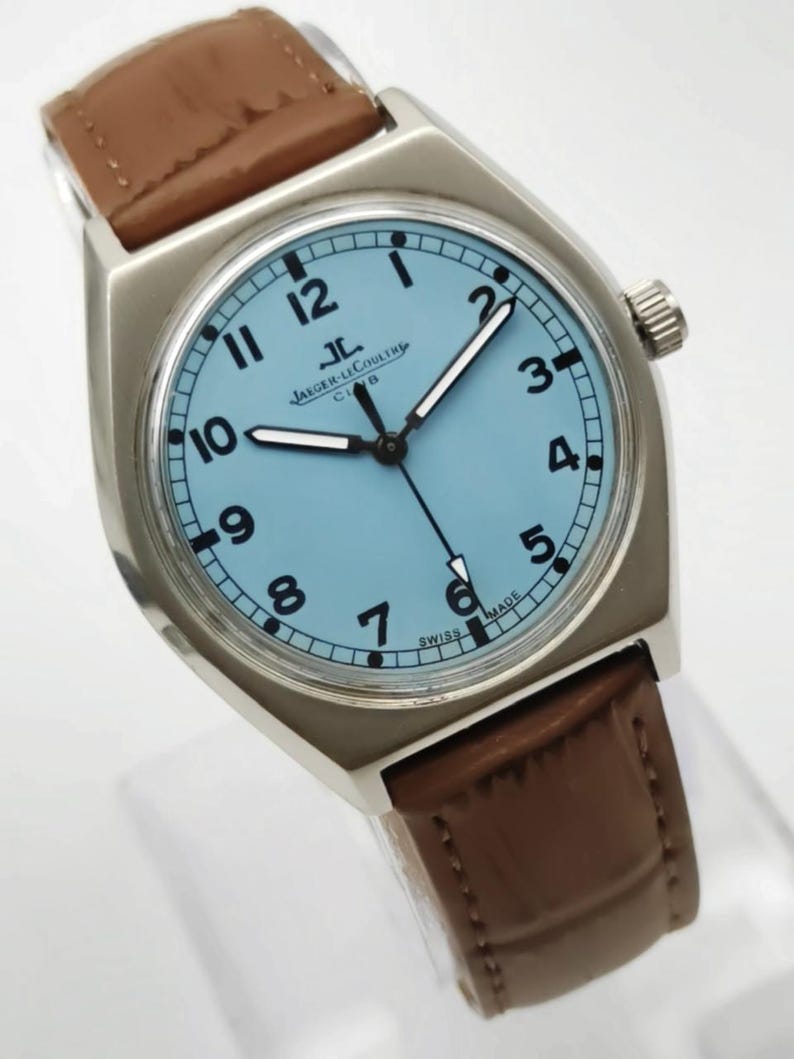 K&ouml;nnte beinhalten: Eine silberfarbene Armbanduhr mit hellblauem Zifferblatt und schwarzen Ziffern. Die Uhr hat ein braunes Lederarmband und den Text "JAEGER-LECOULTRE CLUB SWISS MADE". Die Zeiger der Uhr sind schwarz und wei&szlig;.