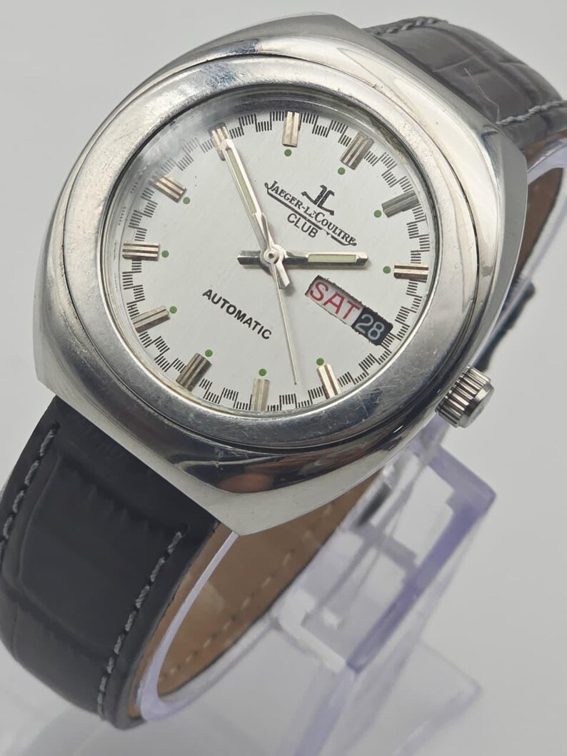 Puede incluir: Un reloj autom&aacute;tico Jaeger-LeCoultre Club plateado con esfera blanca, correa de cuero negro y el d&iacute;a mostrado como S&Aacute;B 28. El reloj tiene un bisel y una corona plateados. El reloj se exhibe sobre un soporte transparente.