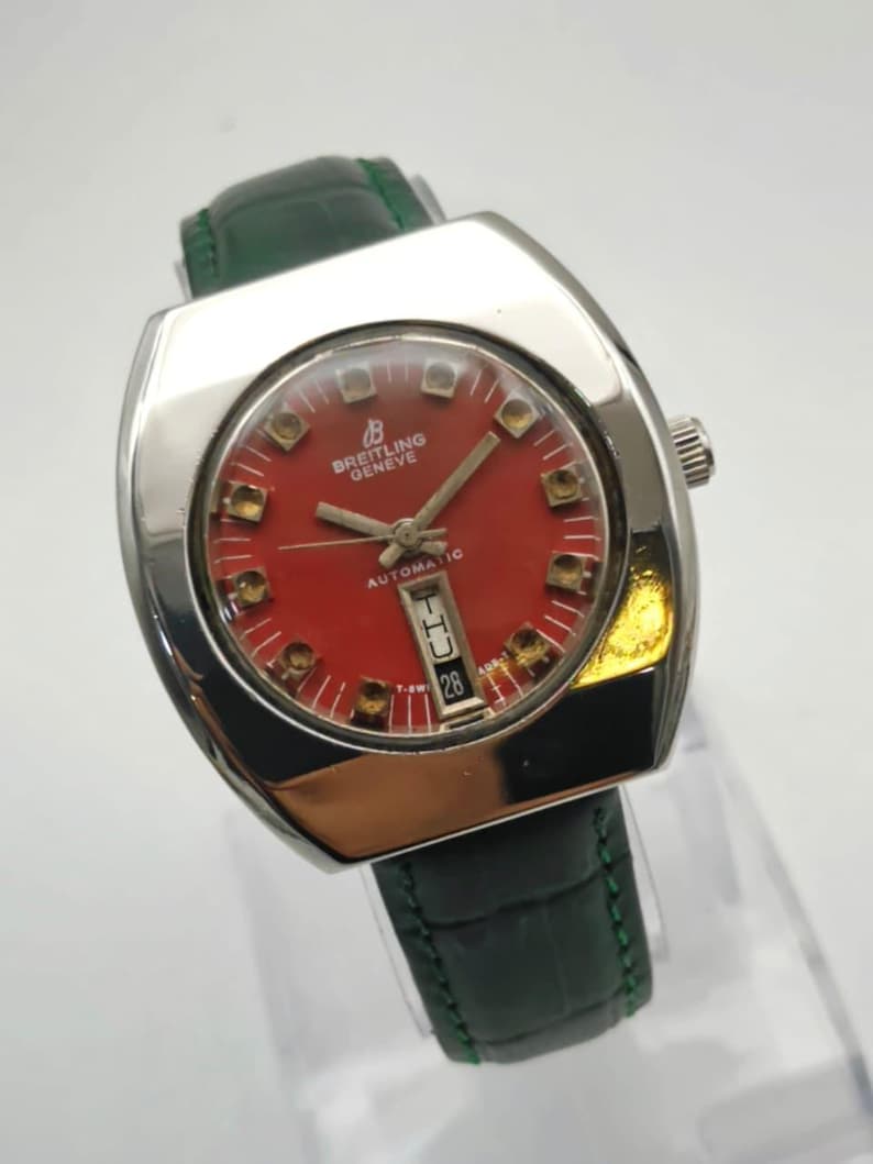 May include: A vintage Breitling Geneve automatic watch with a red dial and a date display. The watch has a silver-tone case and a green leather strap. The watch face features gold-tone hour markers and hands.