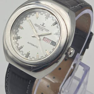 Puede incluir: Un reloj autom&aacute;tico Jaeger-LeCoultre Club plateado con esfera blanca, correa de cuero negro y visualizaci&oacute;n del d&iacute;a y la fecha. El reloj tiene una esfera redonda con agujas y marcadores plateados. La correa mide unos 20 cm.