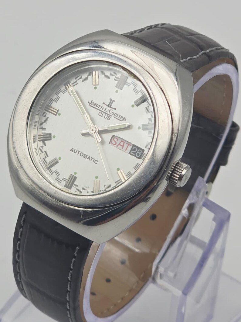 Puede incluir: Un reloj autom&aacute;tico Jaeger-LeCoultre Club plateado con esfera blanca, correa de cuero negro y visualizaci&oacute;n del d&iacute;a y la fecha. El reloj tiene una esfera redonda con agujas y marcadores plateados. La correa mide unos 20 cm.