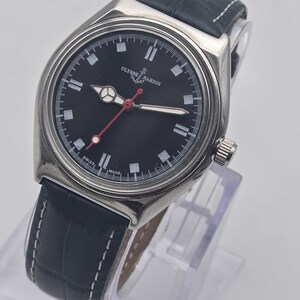 Puede incluir: Un reloj Ulysse Nardin de acero inoxidable con esfera negra, marcadores blancos y segundero rojo. El reloj tiene una correa de cuero negro con costuras blancas. El reloj se exhibe sobre un soporte transparente.