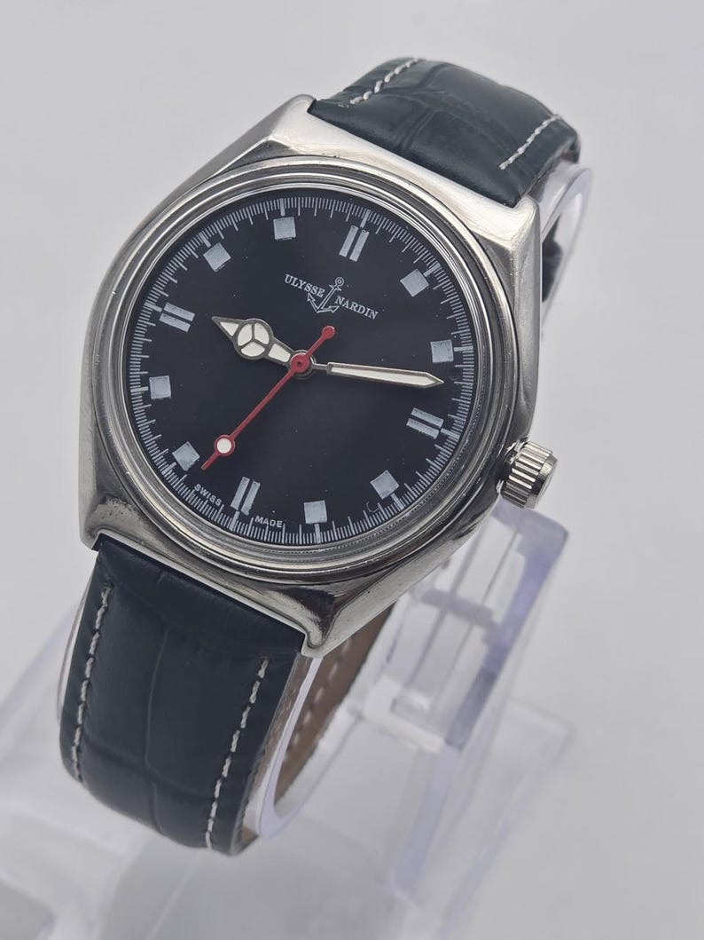 Puede incluir: Un reloj Ulysse Nardin de acero inoxidable con esfera negra, marcadores blancos y segundero rojo. El reloj tiene una correa de cuero negro con costuras blancas. El reloj se exhibe sobre un soporte transparente.