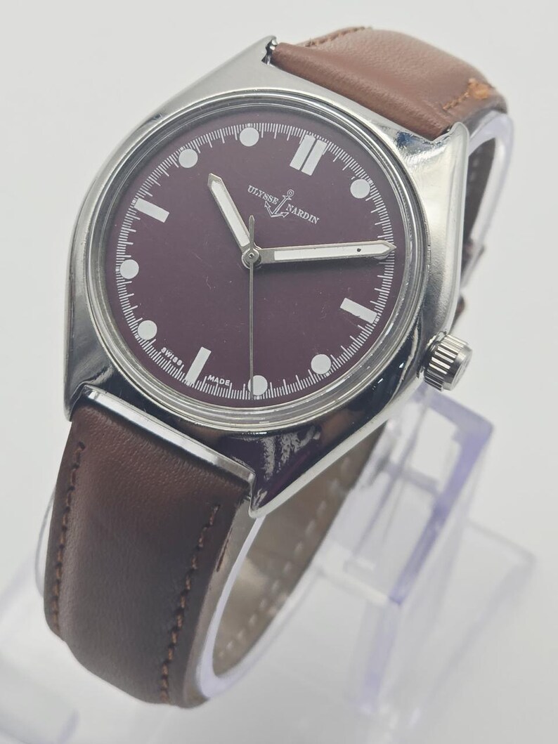 May include: A wristwatch with a burgundy face and a brown leather band. The watch has silver-colored hands and hour markers. The brand name "ULYSSE NARDIN" is displayed on the watch face. The watch is displayed on a clear stand.
