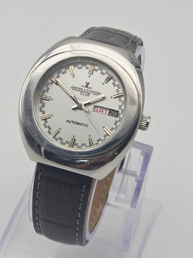 Puede incluir: Un reloj autom&aacute;tico Jaeger-LeCoultre Club plateado con esfera blanca, correa de cuero negra y la fecha mostrada como S&Aacute;B 28. El reloj tiene una esfera redonda y un bisel plateado. La correa es negra con costuras blancas.