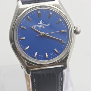 May include: A silver-toned Jaeger-LeCoultre Club watch with a blue dial and a black leather strap. The watch features silver hands and markers, and the words "SWISS MADE" are visible on the dial. The strap is approximately 8 inches long.