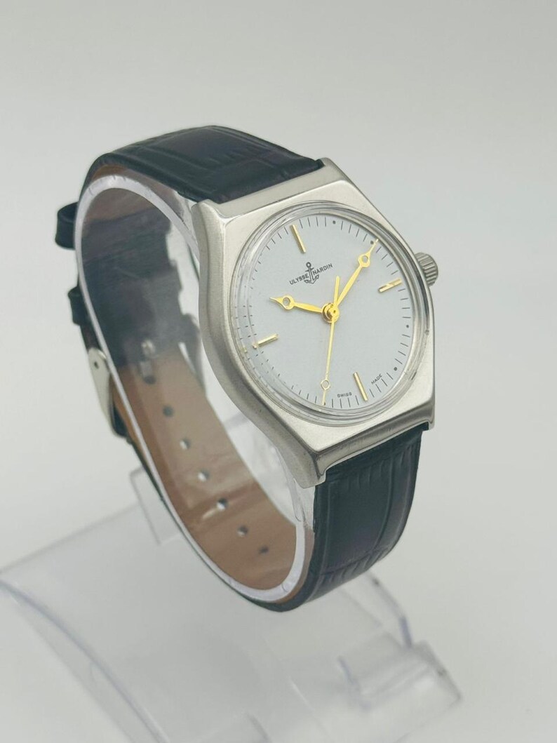 May include: A silver-toned wristwatch with a white face and gold hands and markers. The watch has a black leather band. The watch face has the words "ULYSSSE NARDIN" printed on it.