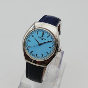 May include: A vintage Jaeger-LeCoultre Club watch with a light blue dial and silver case. The watch has a dark blue leather strap. The watch face displays the Jaeger-LeCoultre logo and text. The watch is on a clear stand.