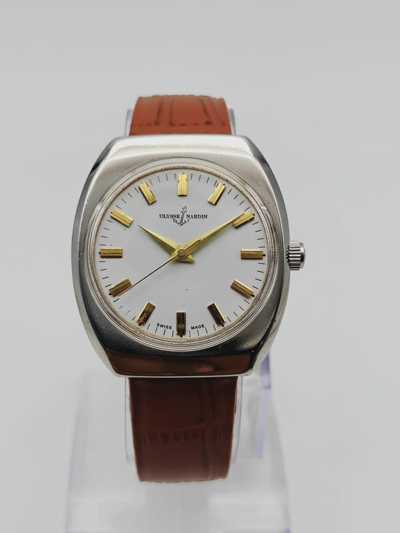 May include: A vintage Ulysse Nardin wristwatch with a silver-toned case and a white dial. The watch features gold-colored hands and markers, and a brown leather strap. The watch is approximately 1.5 inches in diameter.