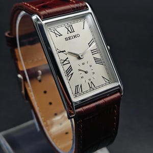 May include: A rectangular Seiko wristwatch with a silver case and a cream-colored face featuring Roman numeral hour markers. The watch has a brown leather band. The watch face displays the brand name and two smaller dials.
