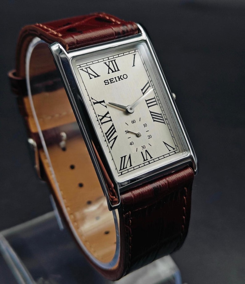 May include: A rectangular Seiko wristwatch with a silver case and a cream-colored face featuring Roman numeral hour markers. The watch has a brown leather band. The watch face displays the brand name and two smaller dials.