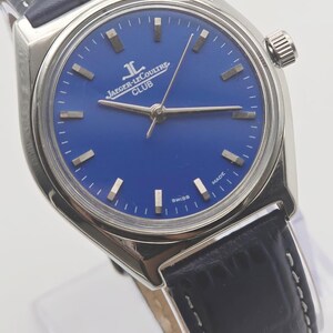 May include: A silver-toned wristwatch with a blue face and a black leather band. The watch face features silver-toned markers and hands, with the text "JAEGER-LECOULTRE CLUB" and "SWISS MADE". The band is dark blue with white stitching.