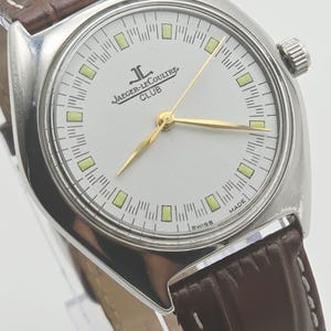 May include: A silver-toned Jaeger-LeCoultre Club watch with a white face, gold hands, and a brown leather band. The watch face features the Jaeger-LeCoultre logo and the word "CLUB". The watch has a round face and a textured brown leather band.