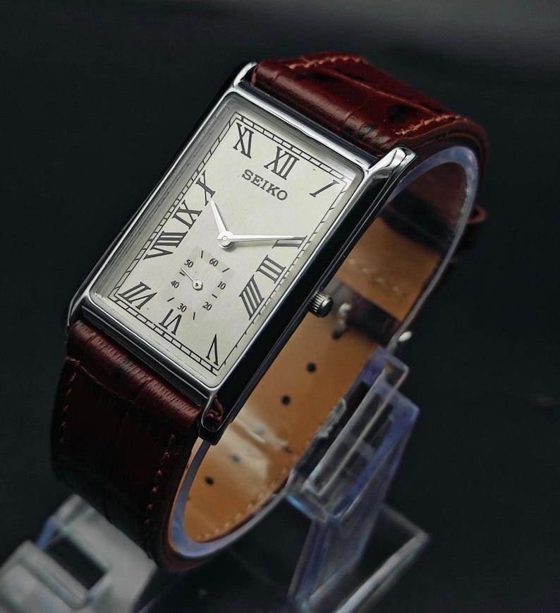 May include: A rectangular Seiko watch with a silver case and a cream-colored face featuring Roman numerals. The watch has a brown leather band. The watch is displayed on a clear stand.