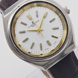 Pu&ograve; includere: Un orologio da polso vintage argentato con quadrante bianco e dettagli dorati. L'orologio presenta un cinturino in pelle marrone e il marchio "Ulysse Nardin" &egrave; visibile. Le lancette e gli indici sono dorati. Fatto in Svizzera.