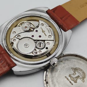 May include: Close-up of a vintage wristwatch with the back cover removed, revealing the intricate mechanical movement. The watch has a silver-colored case and a brown leather strap. The watch mechanism is labeled "Ulysse Nardin".