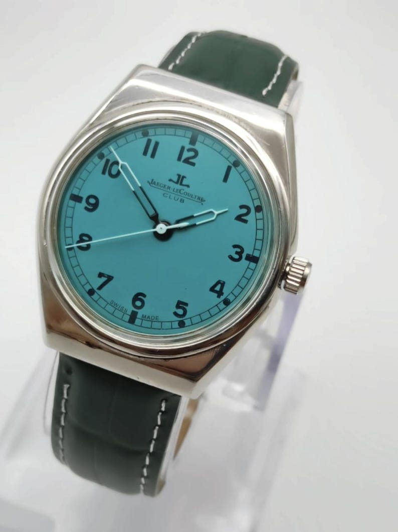 Pode incluir: Um rel&oacute;gio prateado com mostrador turquesa e pulseira de couro verde escuro. O mostrador exibe o logotipo Jaeger-LeCoultre e a palavra "CLUB". A pulseira tem costuras brancas.