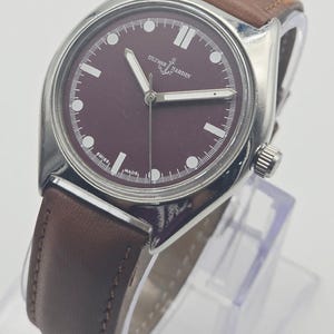 May include: A silver-toned wristwatch with a burgundy face and brown leather band. The watch face features white hour markers and hands, with the brand name "ULYSSE NARDIN" displayed. The watch is displayed on a clear stand.