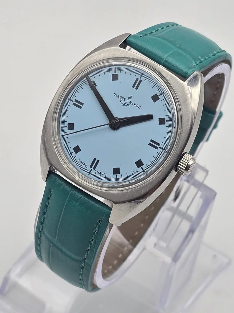 Peut inclure: Une montre Ulysse Nardin vintage avec un cadran bleu clair et des aiguilles noires. La montre a un bo&icirc;tier argent&eacute; et un bracelet en cuir vert d'eau. La montre est pr&eacute;sent&eacute;e sur un support transparent.