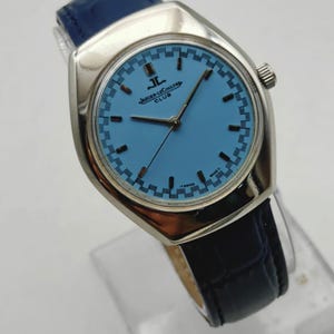 May include: A silver Jaeger-LeCoultre Club watch with a light blue face and a dark blue leather strap. The watch face has a checkered pattern around the edge and the brand name is visible. A classic timepiece.