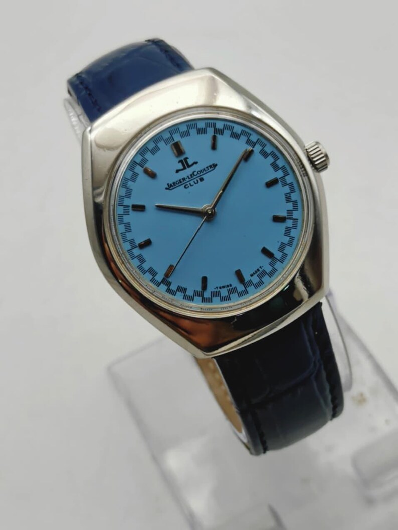 May include: A silver Jaeger-LeCoultre Club watch with a light blue face and a dark blue leather strap. The watch face has a checkered pattern around the edge and the brand name is visible. A classic timepiece.