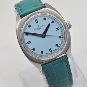 Peut inclure: Une montre-bracelet Ulysse Nardin argent&eacute;e avec un cadran bleu clair et des aiguilles noires. La montre a un bracelet en cuir vert d'eau. Le cadran comporte des index noirs rectangulaires et les mots "Ulysse Nardin" et "Swiss Made".