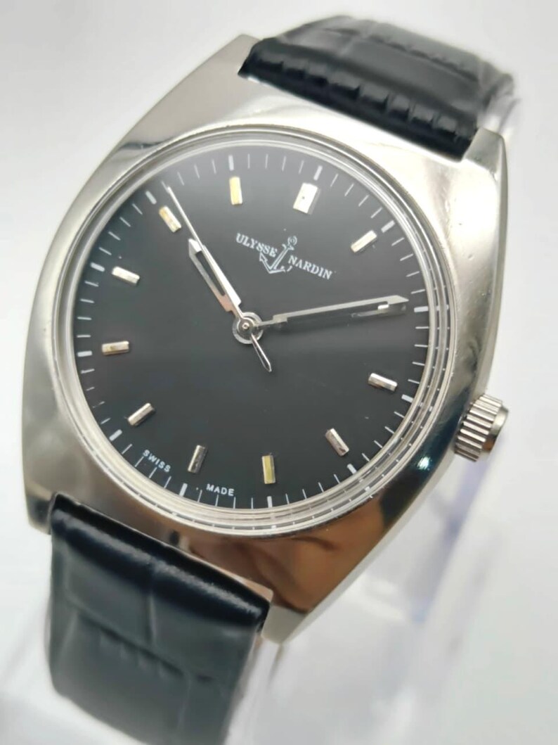 Puede incluir: Un reloj Ulysse Nardin plateado con esfera negra y correa de cuero negro. La esfera del reloj tiene marcadores y agujas plateadas y doradas. El reloj est&aacute; marcado "SWISS MADE".