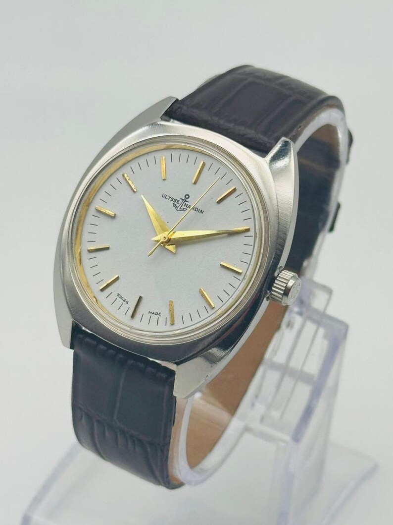 May include: A silver wristwatch with a light-coloured face and gold hands and markers. The watch features the text "ULYSSE NARDIN" and a dark brown leather strap. The watch is displayed on a clear stand.