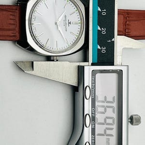 May include: A silver-toned Jaeger-LeCoultre Club watch with a light-colored face and brown leather band. The watch is being measured with a digital caliper, showing a width of 36.94 mm.