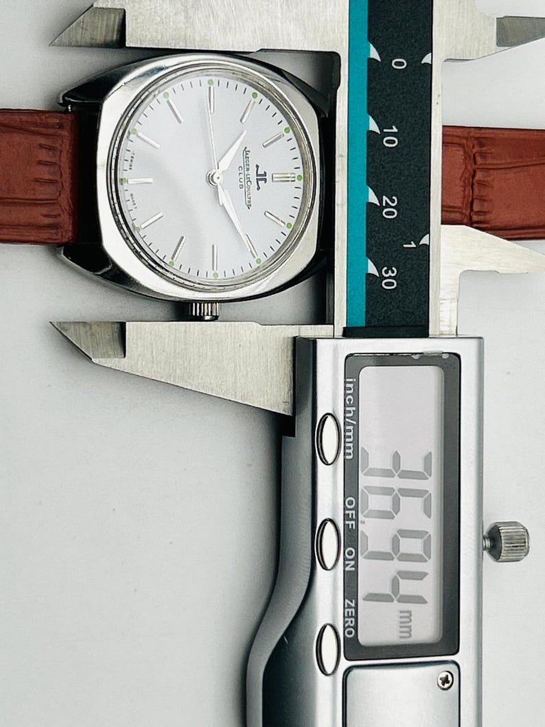 May include: A silver-toned Jaeger-LeCoultre Club watch with a light-colored face and brown leather band. The watch is being measured with a digital caliper, showing a width of 36.94 mm.