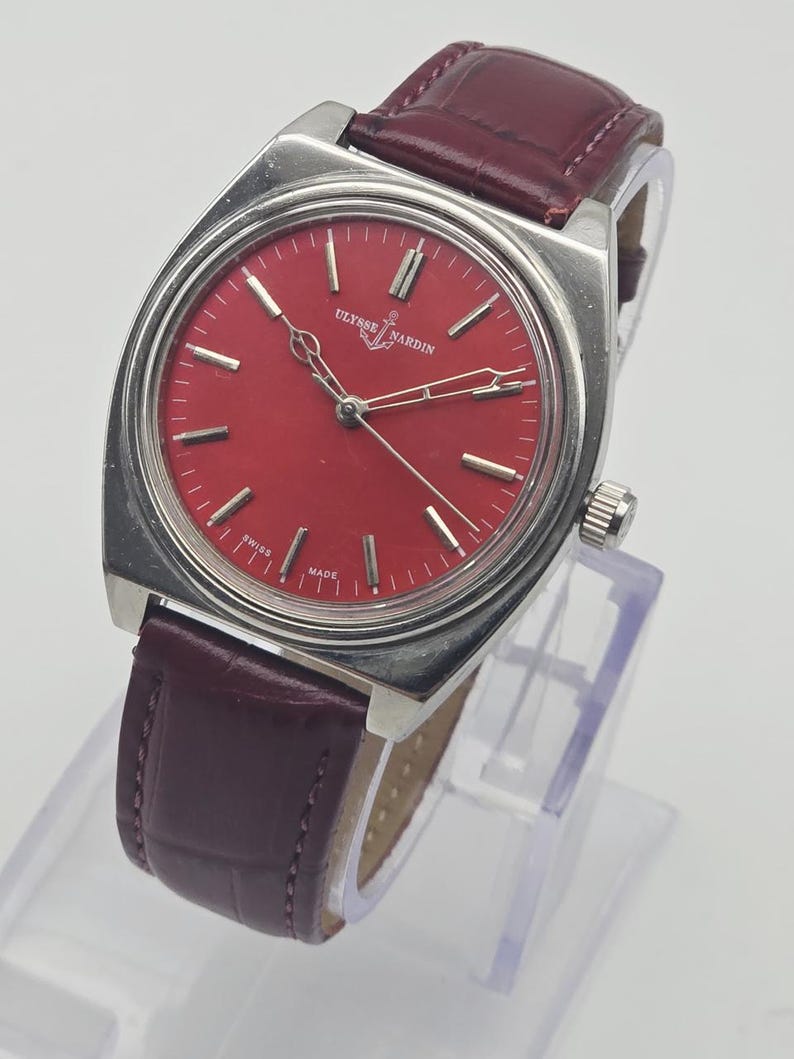 May include: A vintage Ulysse Nardin wristwatch with a red dial and silver-tone case. The watch features silver hands and markers, and a burgundy leather strap. The watch is displayed on a clear stand. The dial reads "Ulysse Nardin" and "Swiss Made".