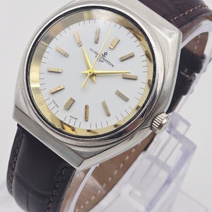 Pu&ograve; includere: Un orologio Ulysse Nardin argentato con quadrante bianco, dettagli dorati e cinturino in pelle marrone. L'orologio ha una forma rettangolare e un cristallo trasparente. Il quadrante mostra il nome del marchio e "SWISS MADE".