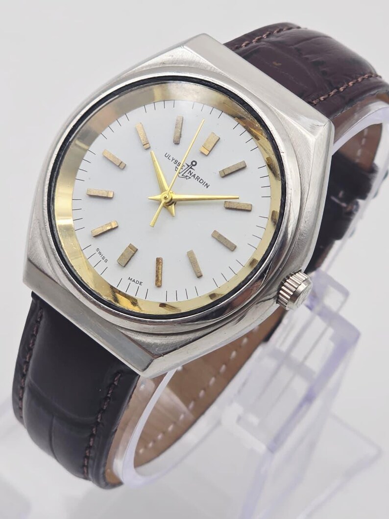 Pu&ograve; includere: Un orologio Ulysse Nardin argentato con quadrante bianco, dettagli dorati e cinturino in pelle marrone. L'orologio ha una forma rettangolare e un cristallo trasparente. Il quadrante mostra il nome del marchio e "SWISS MADE".