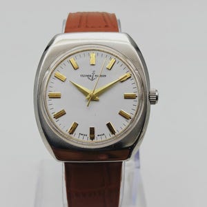 May include: A vintage Ulysse Nardin wristwatch with a silver-toned case and a white dial. The watch features gold-colored hands and markers, and a brown leather strap. The watch is marked "Swiss Made".