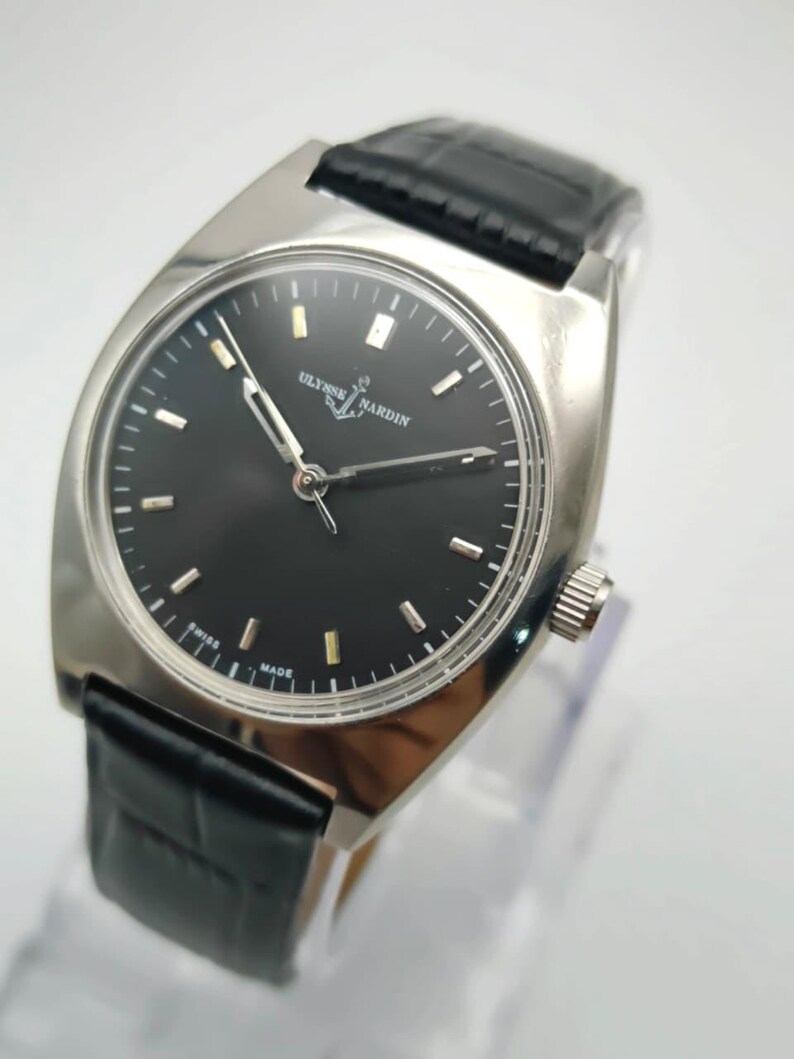 Puede incluir: Un reloj de pulsera plateado con esfera negra y correa de cuero negro. La esfera del reloj presenta marcadores de hora y agujas plateadas. Se muestra la marca "ULYSSE NARDIN". Dise&ntilde;o cl&aacute;sico y elegante.