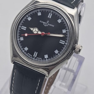 Puede incluir: Un reloj Ulysse Nardin de acero inoxidable con esfera negra y correa de cuero azul oscuro. La esfera presenta marcadores y agujas blancas, con un segundero rojo. La correa est&aacute; cosida con hilo blanco.