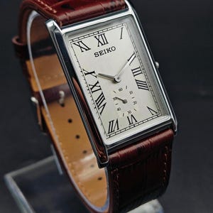 May include: A rectangular Seiko wristwatch with a silver-colored case and a cream-colored face. The watch features black Roman numerals and a small seconds dial. It has a brown leather band.