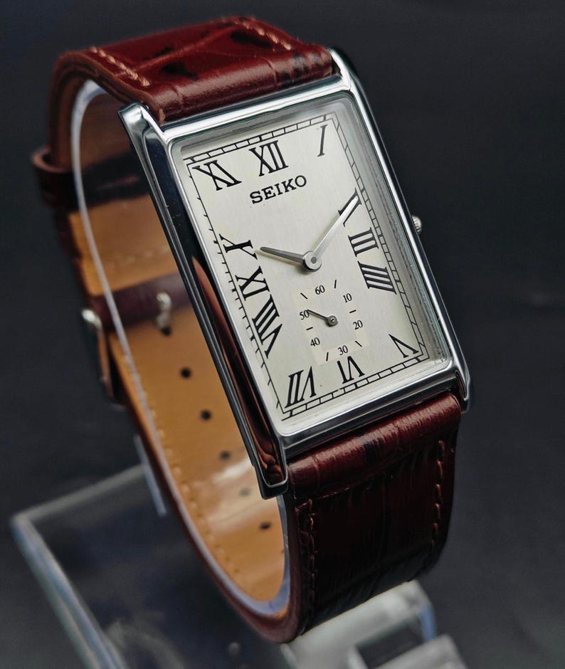 May include: A rectangular Seiko wristwatch with a silver-colored case and a cream-colored face. The watch features black Roman numerals and a small seconds dial. It has a brown leather band.