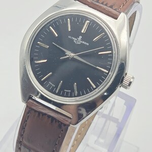 May include: A silver-toned Ulysse Nardin wristwatch with a black face and brown leather band. The watch has silver hands and markers, and the band is a dark brown with a textured pattern. The watch face reads "Ulysse Nardin" and "Swiss Made."