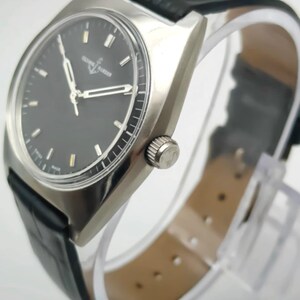 Puede incluir: Un reloj de pulsera plateado con esfera negra y correa de cuero negro. El reloj tiene una caja rectangular y una corona plateada. La esfera tiene marcadores y agujas plateadas. La correa mide unos 20 cm de largo.
