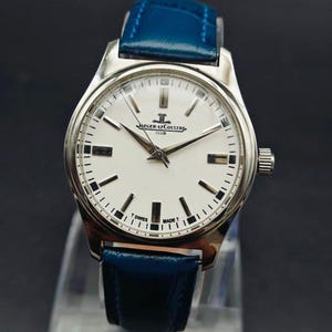 Peut inclure: Une montre Jaeger-LeCoultre Club argent&eacute;e avec un cadran blanc et un bracelet en cuir bleu. Le cadran comporte des marqueurs et des aiguilles noirs, avec le nom de la marque et le texte "T SWISS MADE T". Le bracelet est bleu vif.