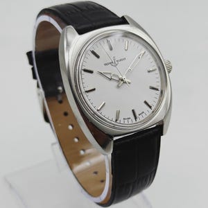 May include: A silver-toned wristwatch with a white face and black leather band. The watch features the text "ULYSSES NARDIN SWISS MADE" on the face. The band has a crocodile pattern.