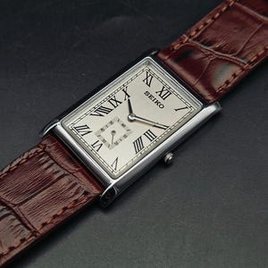 May include: A rectangular Seiko watch with a silver case and a cream-colored face featuring Roman numerals. The watch has a brown leather band. The watch face displays the brand name and two smaller dials.