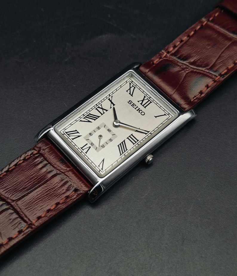 May include: A rectangular Seiko watch with a silver case and a cream-colored face featuring Roman numerals. The watch has a brown leather band. The watch face displays the brand name and two smaller dials.
