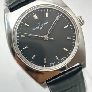 Puede incluir: Un reloj de pulsera Ulysse Nardin plateado con esfera negra y correa de cuero negro. El reloj tiene una esfera redonda con marcadores plateados y dorados. La esfera tiene las palabras "Ulysse Nardin" y "Swiss Made".