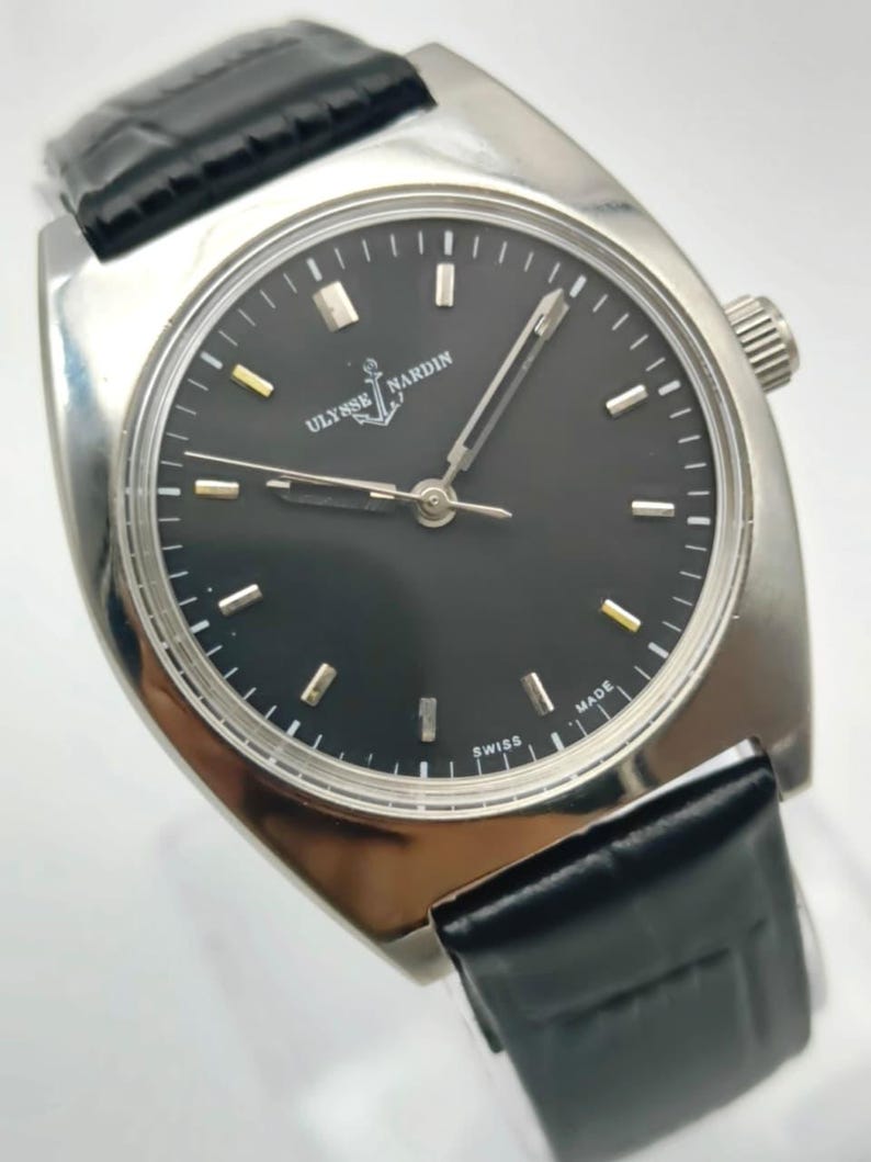 Puede incluir: Un reloj de pulsera Ulysse Nardin plateado con esfera negra y correa de cuero negro. El reloj tiene una esfera redonda con marcadores plateados y dorados. La esfera tiene las palabras "Ulysse Nardin" y "Swiss Made".