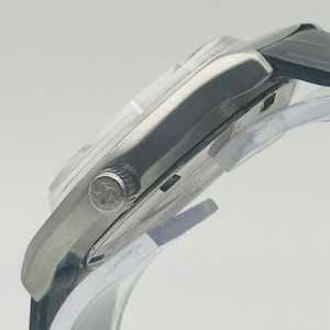 May include: A silver-toned wristwatch with a black leather band. The watch face is not visible, but the side profile shows a clear crystal and a textured crown. The watch is displayed on a clear stand.